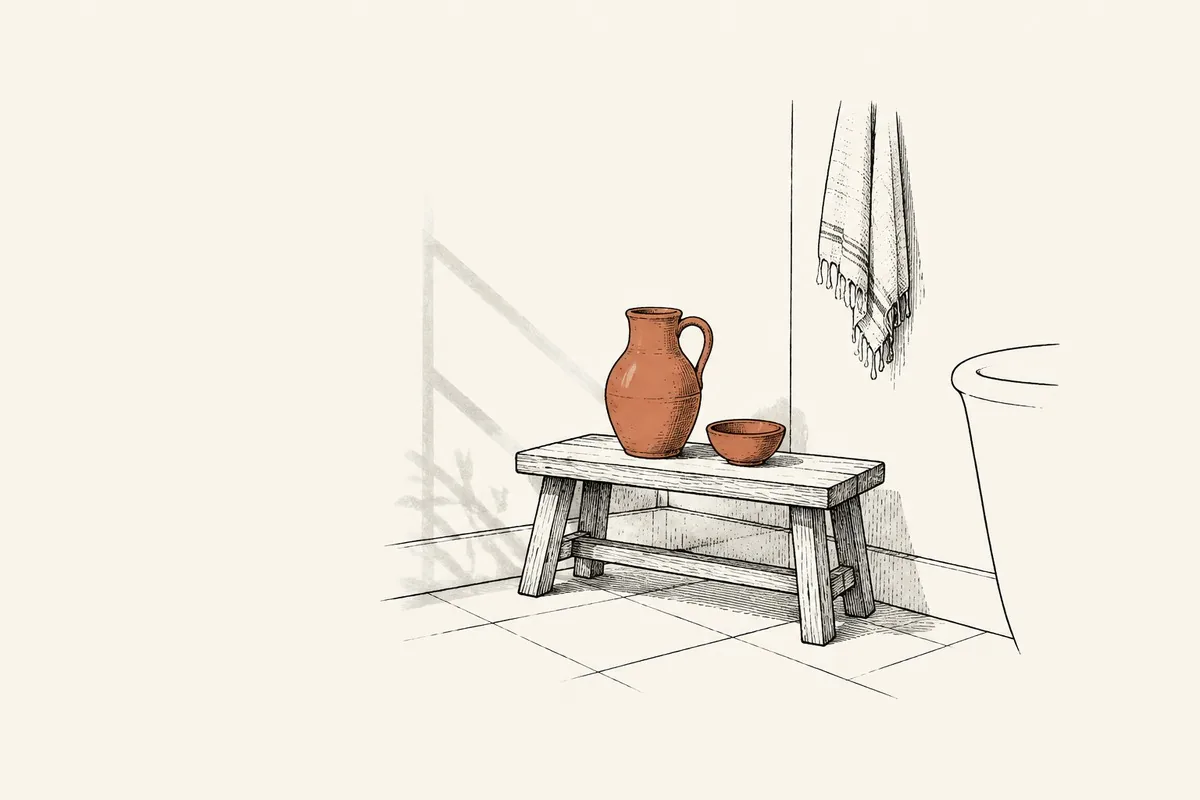 Un banco bajo de madera en un rincón del baño, con una jarra de cerámica y un cuenco pequeño encima, una toalla clara colgada al lado y el borde de una pila asomando por la derecha, y la luz de la mañana entrando de lado